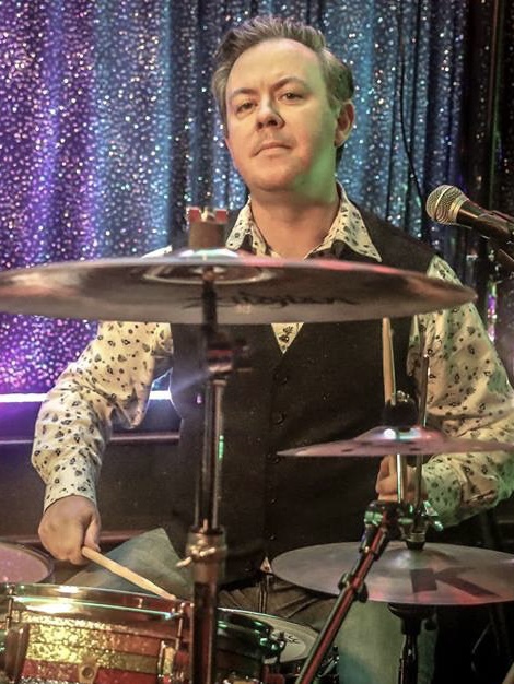 IndieFly drummer Dave Thorbinson sat at the drums during a gig, wearing a white shirt and grey waistecoat.