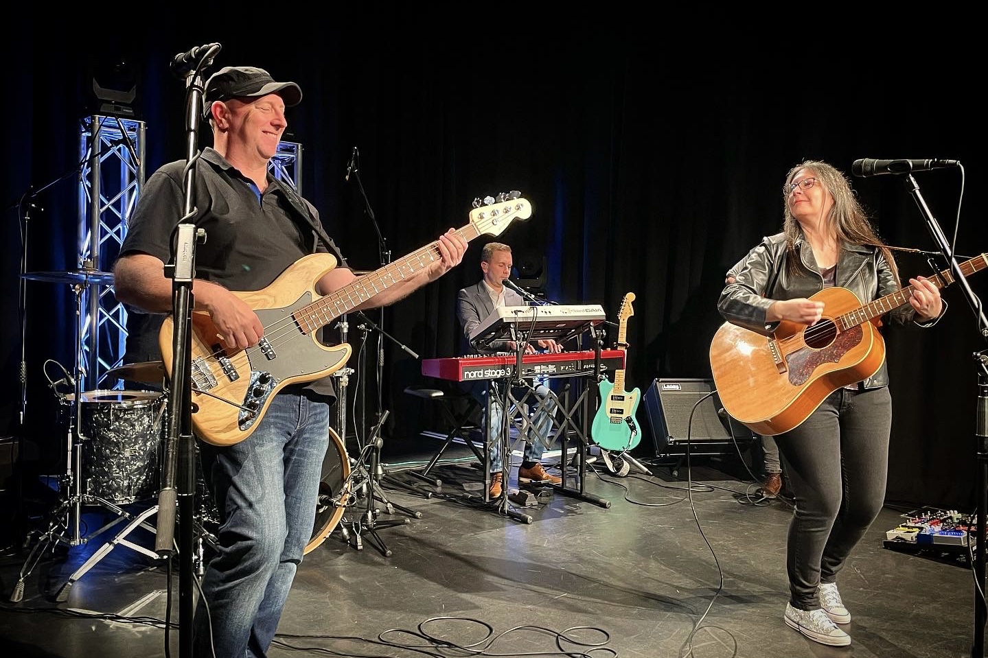 indie band IndieFly performing on stage: singer Emma and bassist Moxy smile at each other while playing guitars, with Craig playing keyboards in the background.